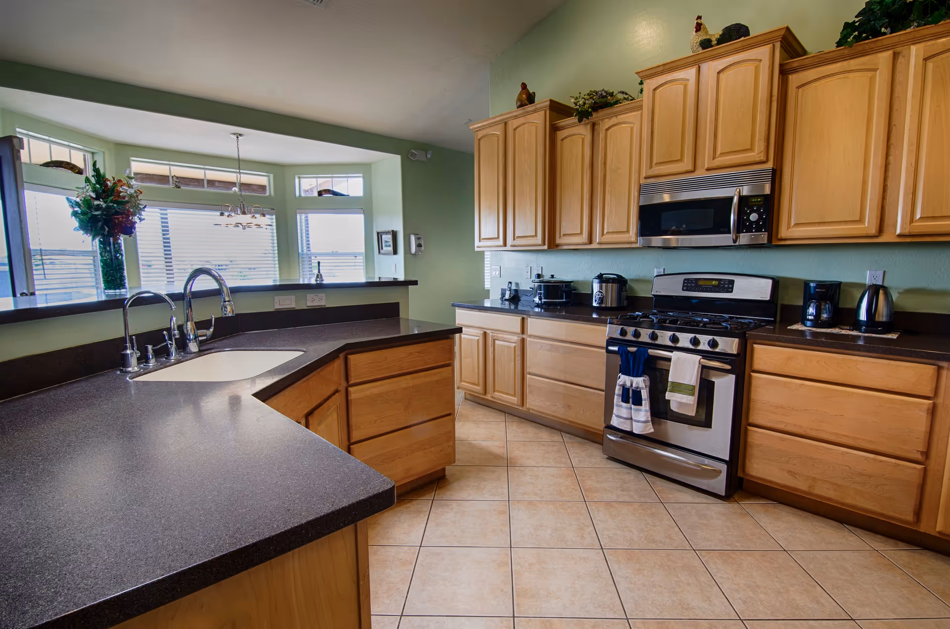A bright kitchen with light wood cabinets, a stainless steel stove and microwave, and a dark countertop with a built-in sink. There is a large window with blinds and a hanging flower pot, and the floor is tiled.