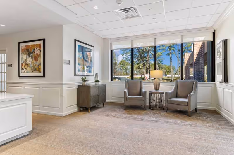 A bright and spacious sitting area in a senior living facility named Leisure Park, featuring two gray armchairs with a small round table and lamp between them, large windows showing trees and a brick building outside, light-colored walls with decorative paneling, framed artwork on the walls, and a carpeted floor.