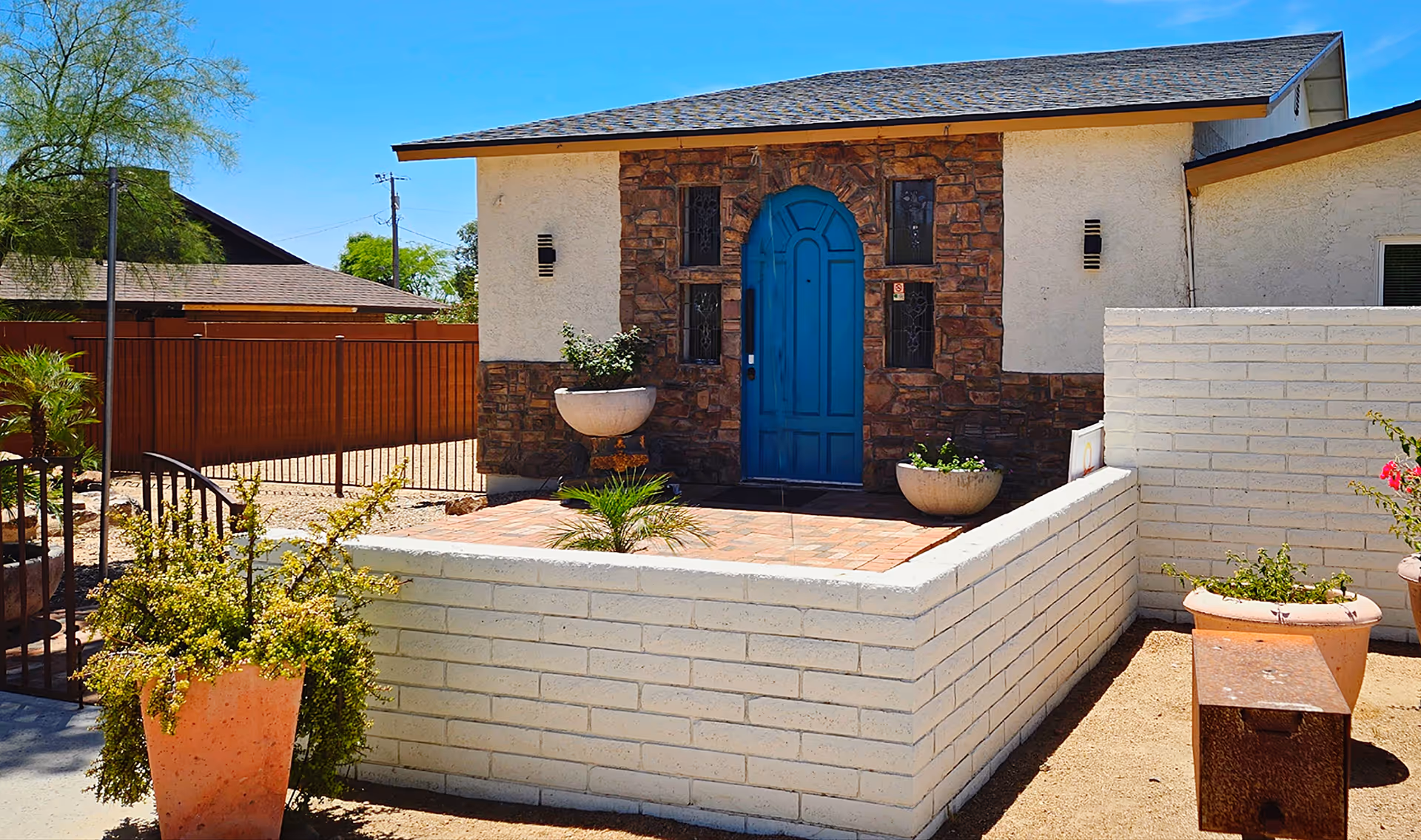 Exterior view of a building with a blue arched door set in a stone facade. The entrance is surrounded by a white brick wall with potted plants on the patio area. There is a wooden fence and some trees in the background under a clear blue sky.