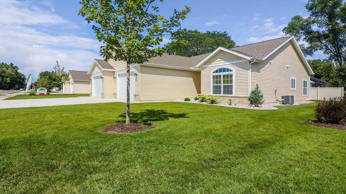 Exterior view of a single-story beige building with a gable roof, surrounded by a well-maintained green lawn and small trees under a partly cloudy sky.