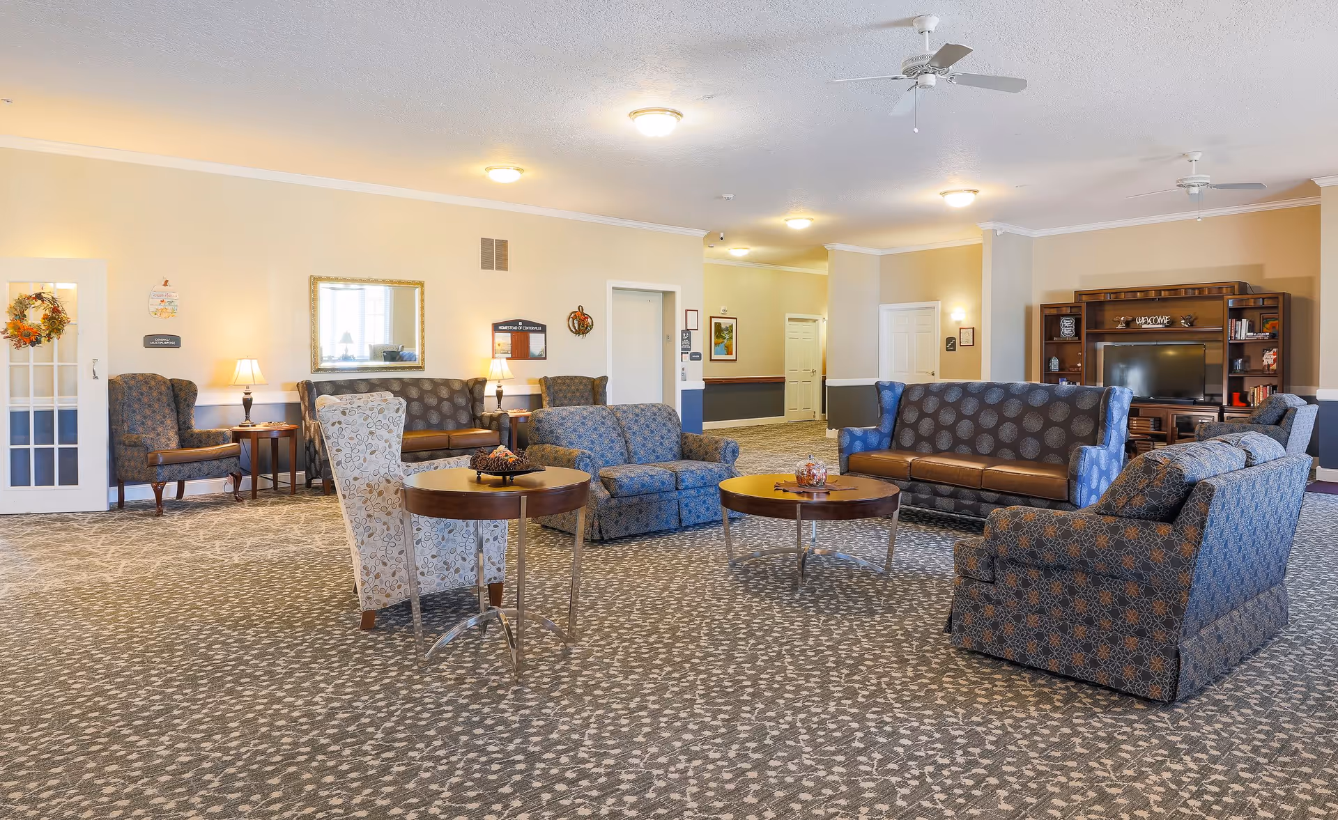 A spacious living room area in an assisted living facility with multiple upholstered sofas and armchairs arranged around two round wooden coffee tables. The room has patterned carpet, beige walls with white trim, ceiling fans, and soft lighting from wall lamps and ceiling fixtures. A large wooden entertainment center with a TV and decorative items is visible in the background.