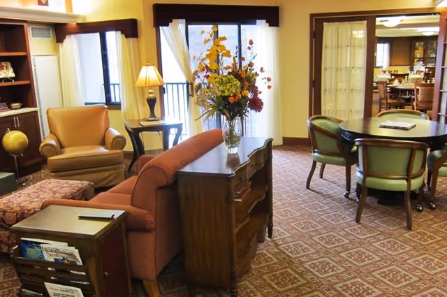 A cozy living room area with a red sofa, a wooden side table with a vase of flowers, an armchair, a patterned ottoman, and a round dining table with four chairs. The room has large windows with sheer curtains allowing natural light to enter, and a carpet with a geometric pattern covers the floor.