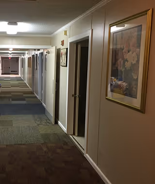 A long indoor hallway in an assisted living facility with multiple doors on the right side and framed artwork hanging on the walls. The floor is covered with a patterned carpet and the ceiling has fluorescent lighting.