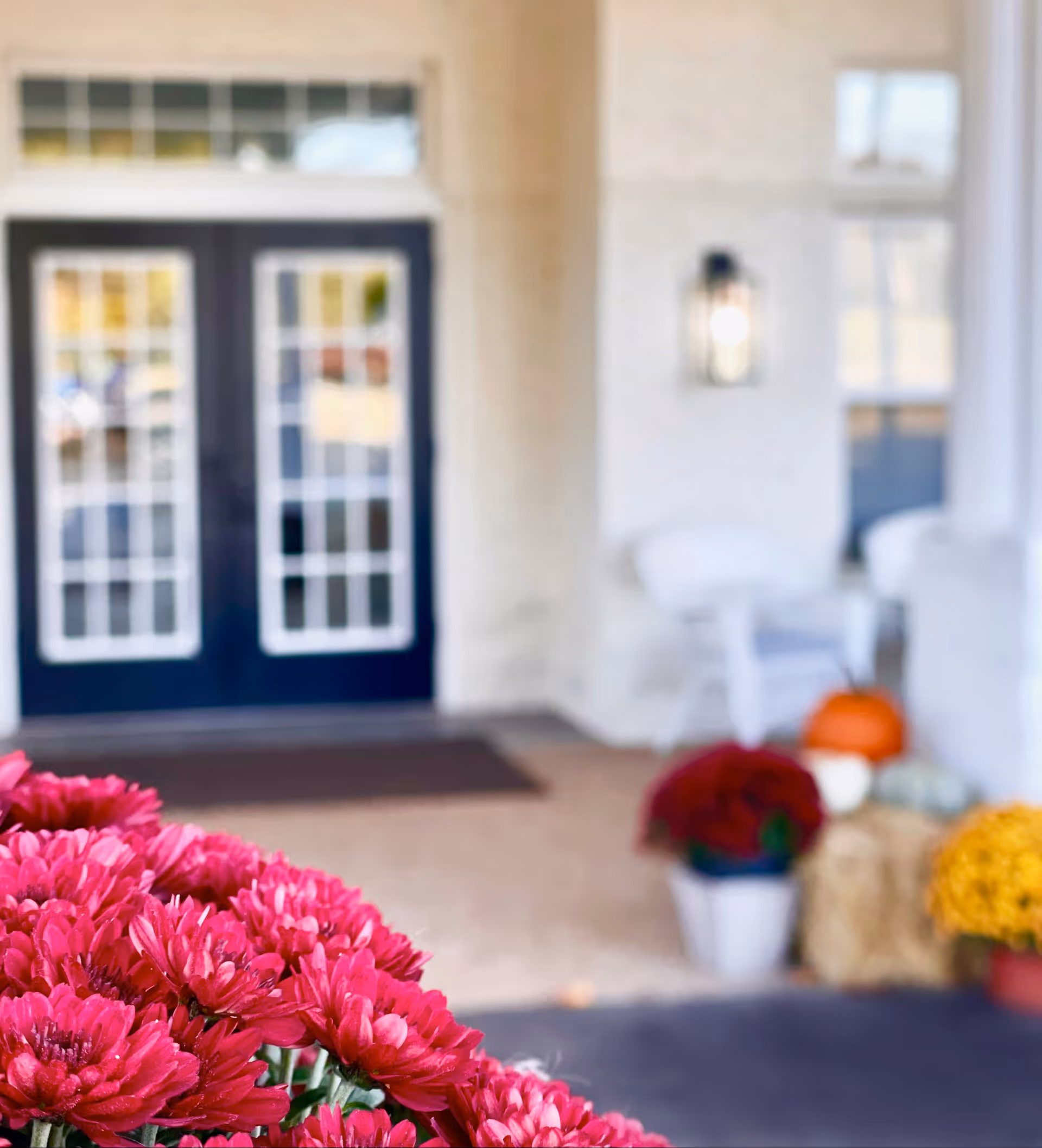 Close-up of vibrant pink flowers in the foreground with a blurred view of a porch entrance featuring double glass doors, white chairs, pumpkins, and potted flowers in the background.