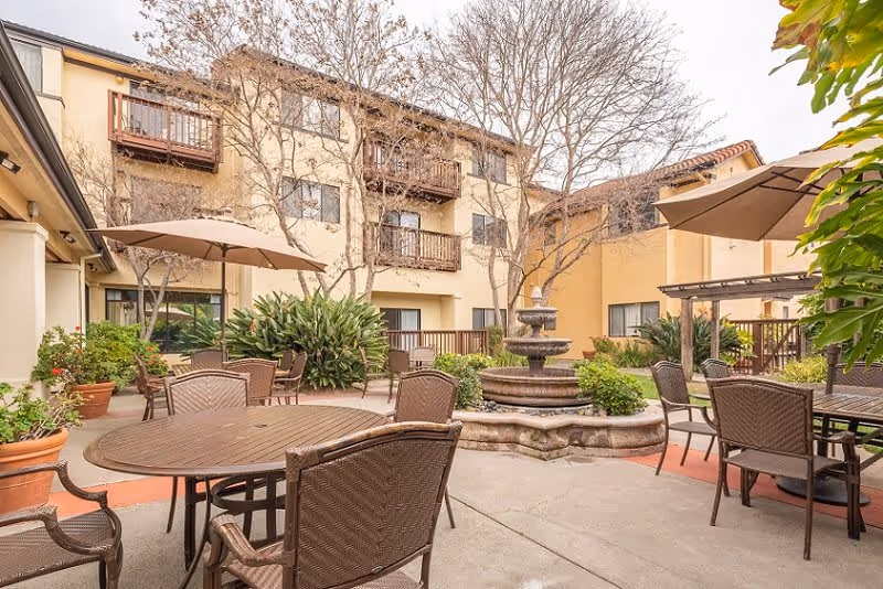 Outdoor courtyard area at Drake Terrace with round tables and chairs, large umbrellas, a central multi-tiered water fountain, potted plants, and a three-story building with balconies in the background.
