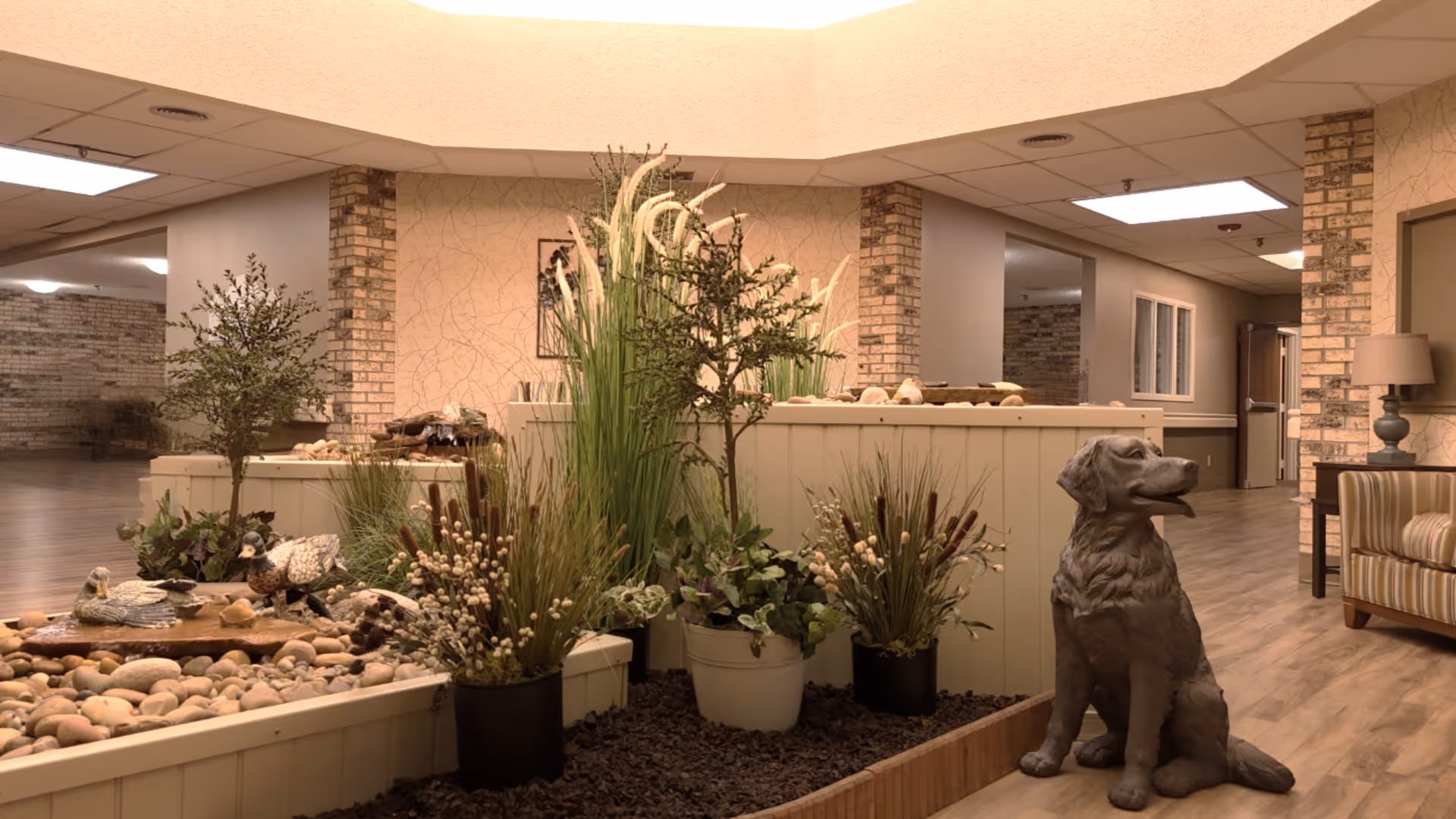 Indoor area of a senior living facility featuring a decorative garden with various plants and a stone water feature surrounded by pebbles. A statue of a sitting dog is placed next to the garden. The space has wood flooring, beige walls with brick accents, and ceiling lights. A striped armchair and a lamp on a small table are visible on the right side.