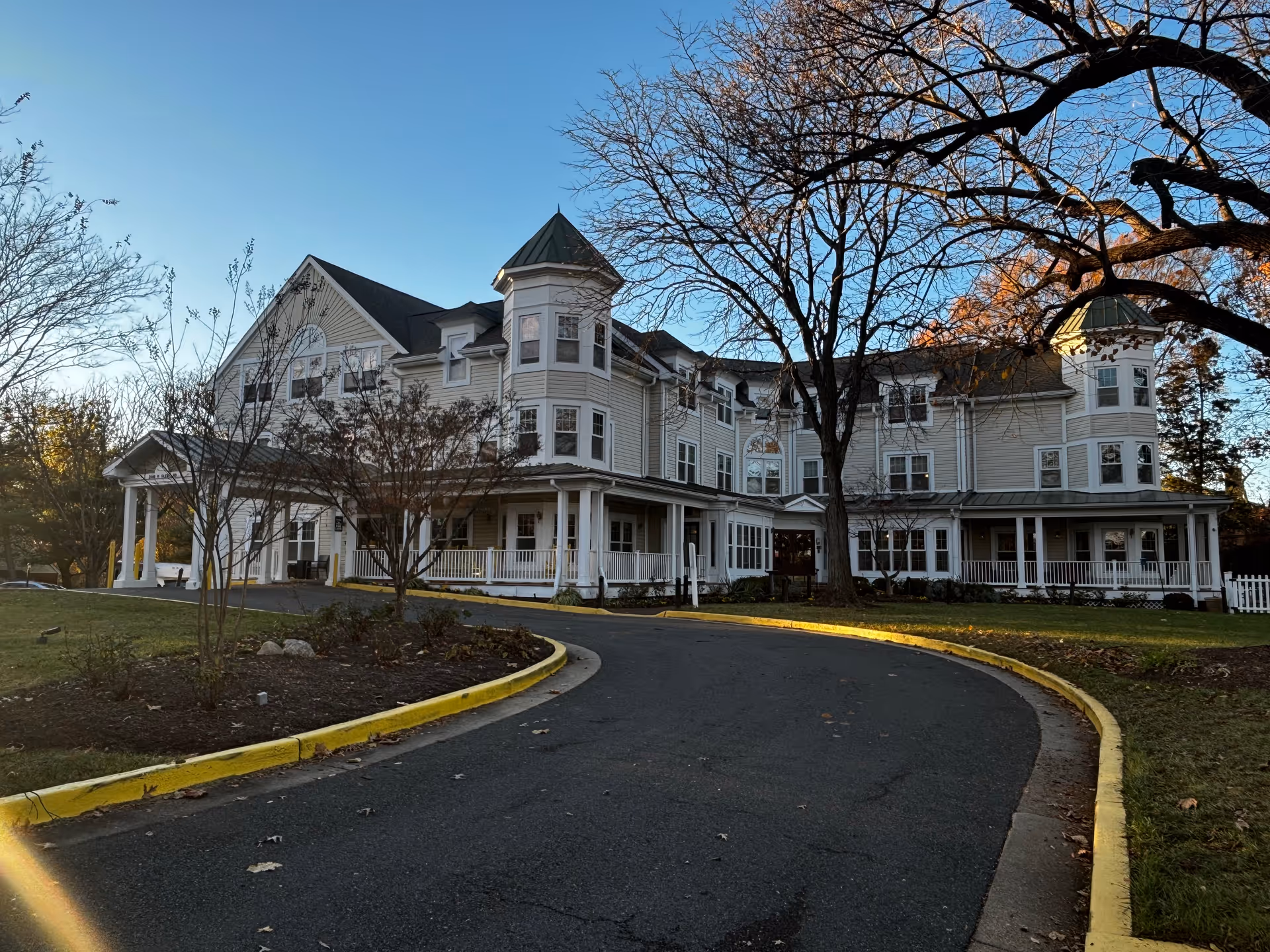 Three-story light-colored Victorian-style building with turrets, a wraparound porch, and a curved driveway bordered by a yellow curb.