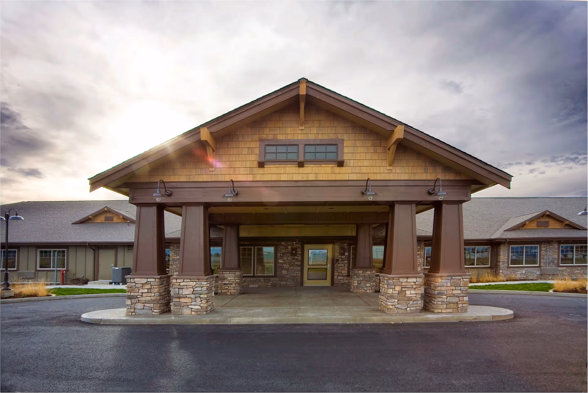 Front exterior view of Creekside Inn Memory Care Community building with a covered entrance supported by stone and wooden pillars, under a cloudy sky.