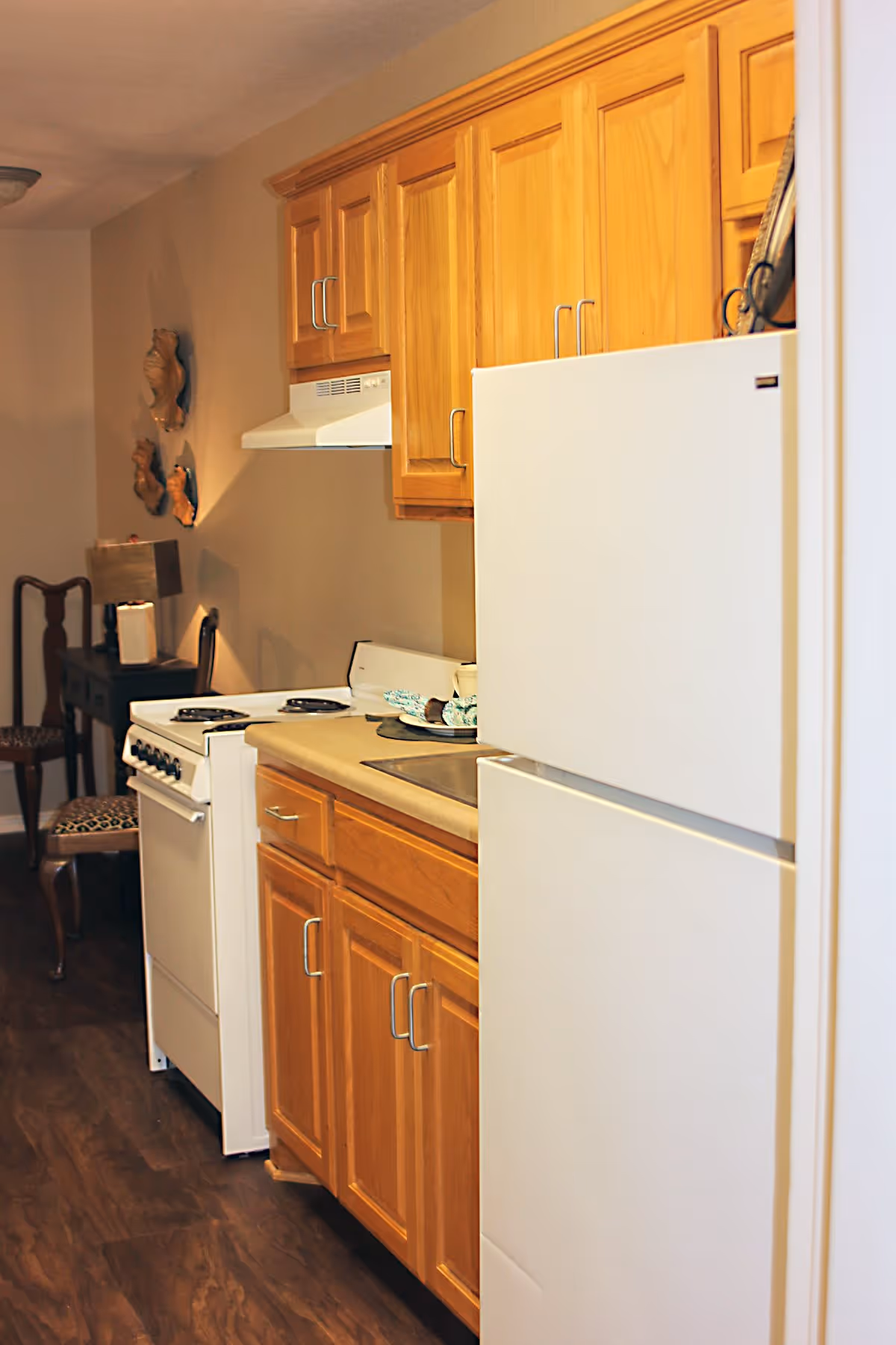 A small kitchen area with wooden cabinets, a white refrigerator, a white stove with an exhaust hood, and a countertop with a sink. In the background, there is a wooden chair with a patterned cushion and a small table with a lamp and decorative wall hangings.