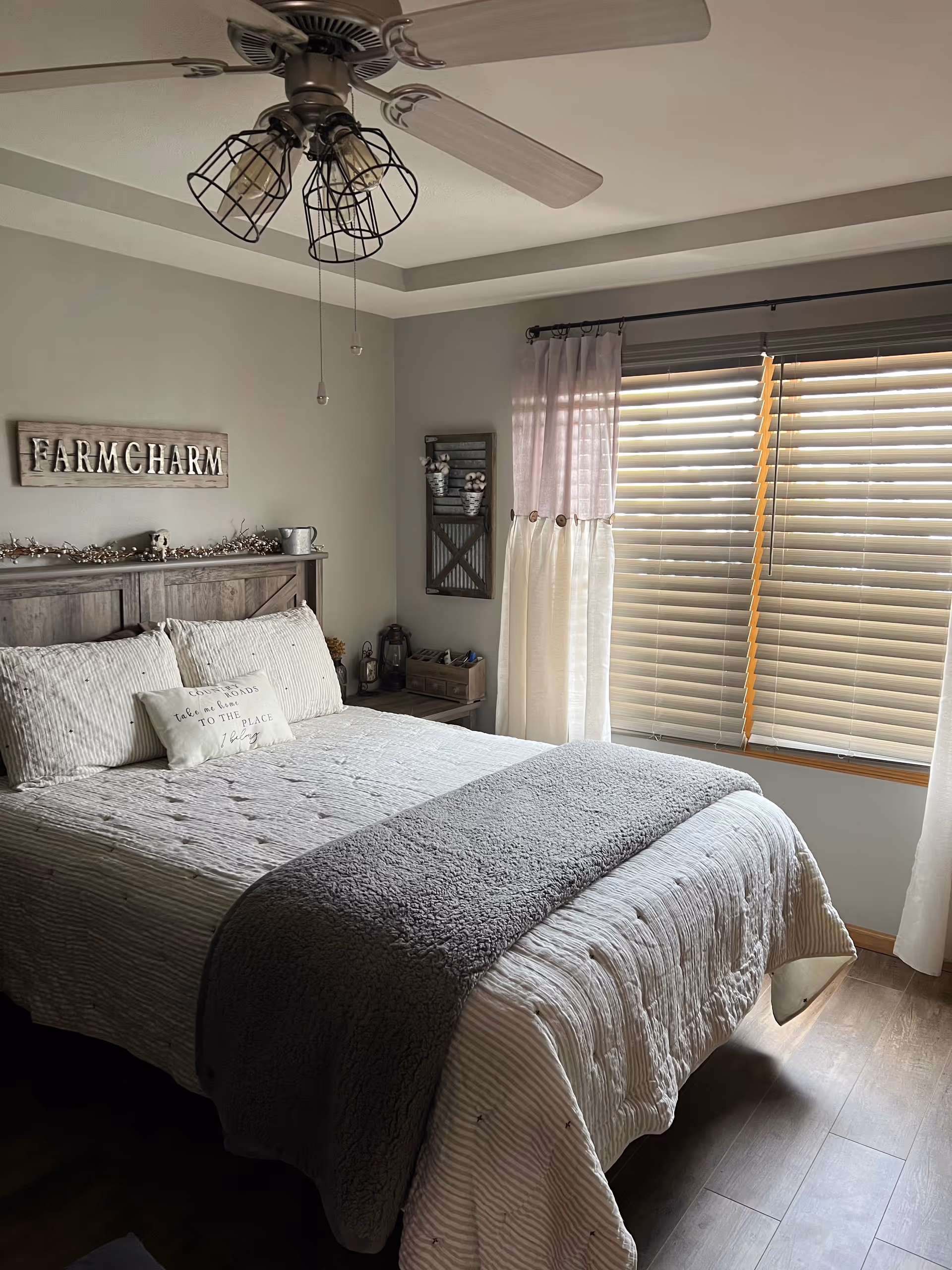 A cozy bedroom with a neatly made bed featuring striped bedding and a gray throw blanket. Above the wooden headboard is a decorative sign that reads 'FARM CHARM'. To the right, there is a window with wooden blinds and two-tone curtains. A ceiling fan with three cage-style light fixtures hangs from the ceiling. The room has light gray walls and wooden flooring.