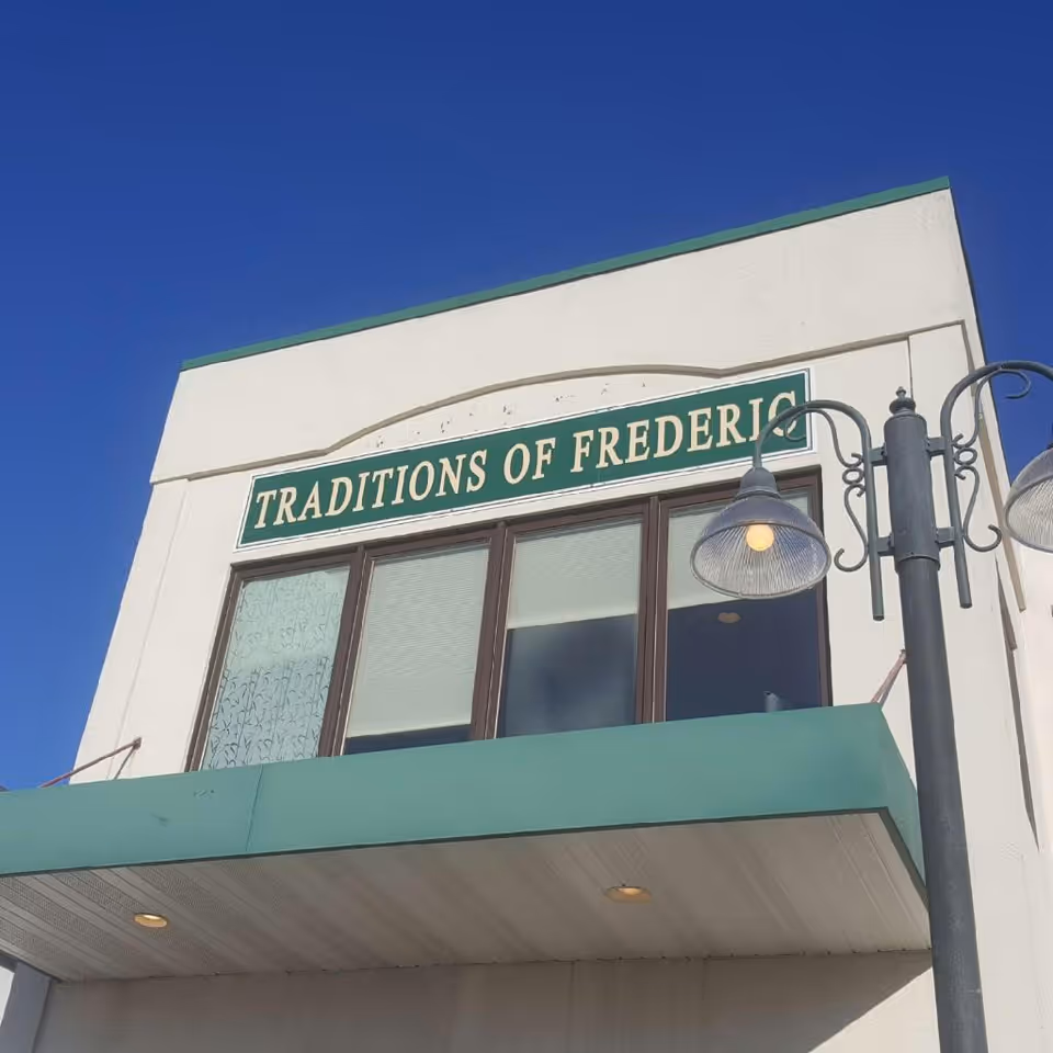 Exterior view of a building with a green sign that reads 'TRADITIONS OF FREDERIC' mounted above three windows. The building has a white facade with a green awning and two decorative street lamps in front. The sky is clear and blue.