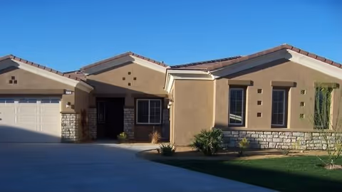 Single-story residential building with beige stucco walls, stone accents, and a tiled roof under a clear blue sky. The house features a two-car garage, multiple windows, and a small landscaped front yard with grass and shrubs.