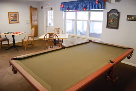 A recreational room with a pool table in the foreground and a table with chairs in the background near a window with blue curtains. There is a dartboard on the wall and a sign that says 'Billiards'.