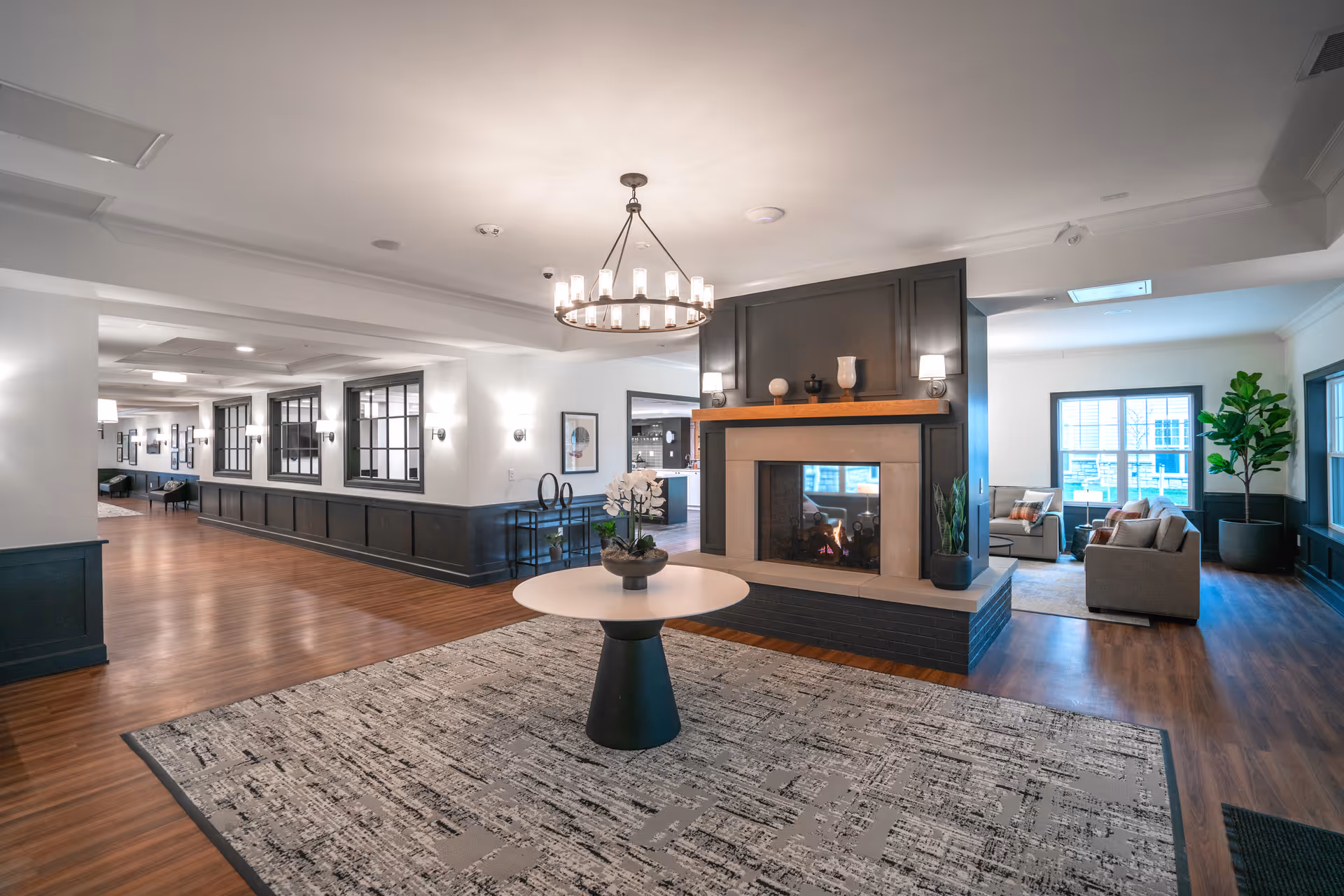 A spacious and well-lit common area in Independence Village of Madison Heights featuring a modern fireplace with decorative items on the mantle, a round table with a flower arrangement in the center, wood flooring, and comfortable seating areas near large windows with natural light.