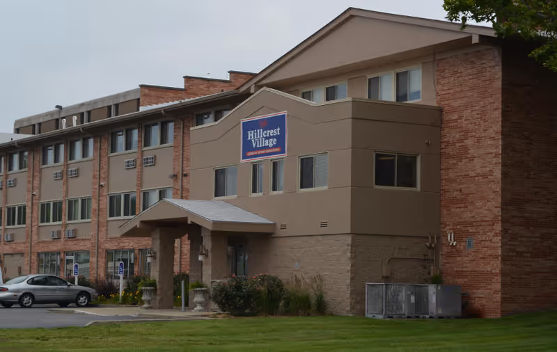 Exterior view of a multi-story senior living facility building with a sign that reads Hillcrest Village. The building has a combination of brick and beige siding, with a covered entrance and several windows. There are a few parked cars and a well-maintained lawn in front.