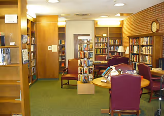 Interior library-style common room with bookshelves, tables, chairs, and a wall clock.