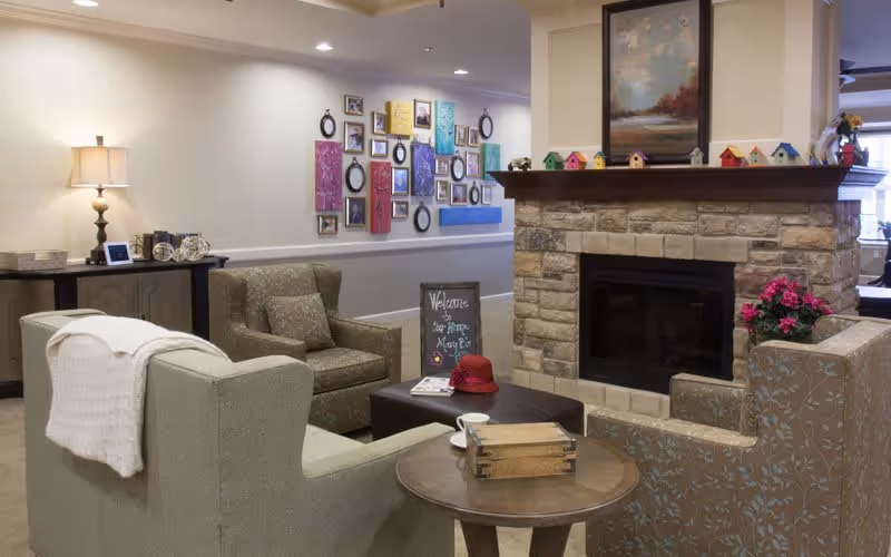 A cozy living room area in a senior living facility with comfortable armchairs and a sofa arranged around a stone fireplace. The fireplace mantel is decorated with small colorful birdhouses and a framed painting above it. A side table holds a wooden box and a cup, while a chalkboard sign on a coffee table reads 'Welcome to Our Home, May 25'. The wall in the background features a collage of framed pictures and artwork. A lamp and decorative items are on a console table against the wall.