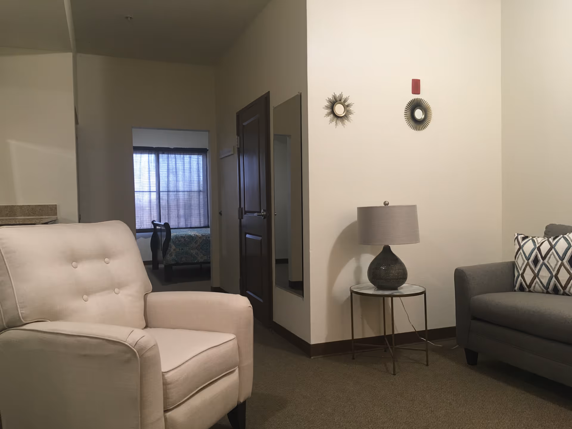 A living room with a beige armchair, gray sofa with patterned pillows, a side table lamp, wall decor, and an open doorway to a bedroom.