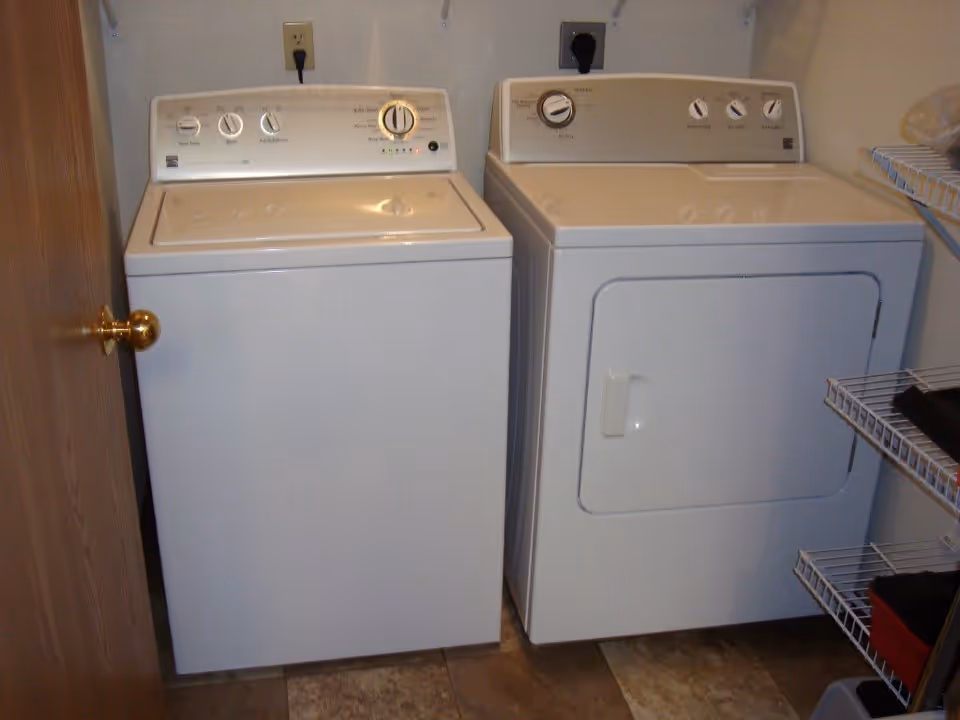 A laundry room with a white top-loading washing machine on the left and a white front-loading dryer on the right. There are wire shelves on the right side holding some folded clothes and other items. The floor has brown and beige tiles, and a wooden door with a brass knob is partially visible on the left.
