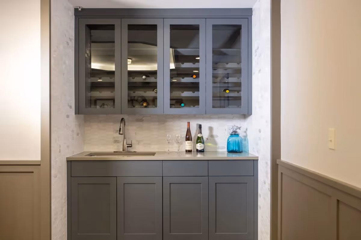 A small kitchenette area with gray cabinets and a countertop. The upper cabinets have glass doors displaying a few wine bottles. On the countertop, there is a sink with a modern faucet, two wine glasses, two bottles of wine, and a blue glass container. The walls are light-colored with a textured tile backsplash behind the sink.