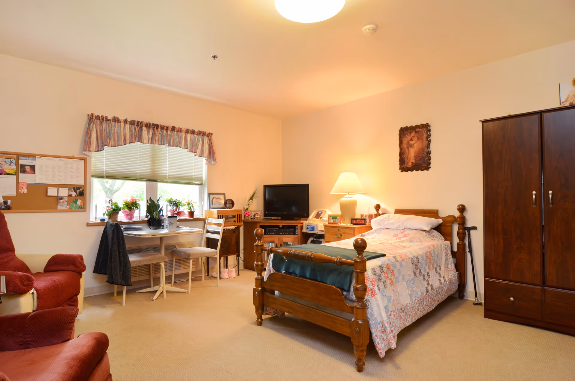 A cozy furnished bedroom with a wooden bed, armchairs, wardrobe, desk by a window and potted plants.