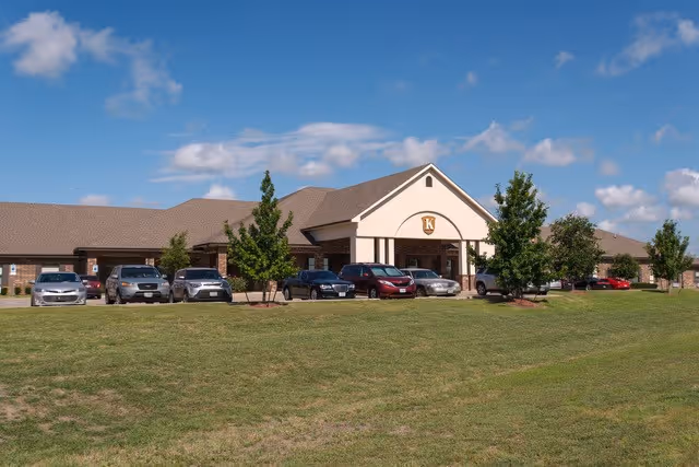 Front exterior view of Kemp Care Center building with a large lawn in the foreground and several parked cars in front of the entrance under a blue sky with scattered clouds.