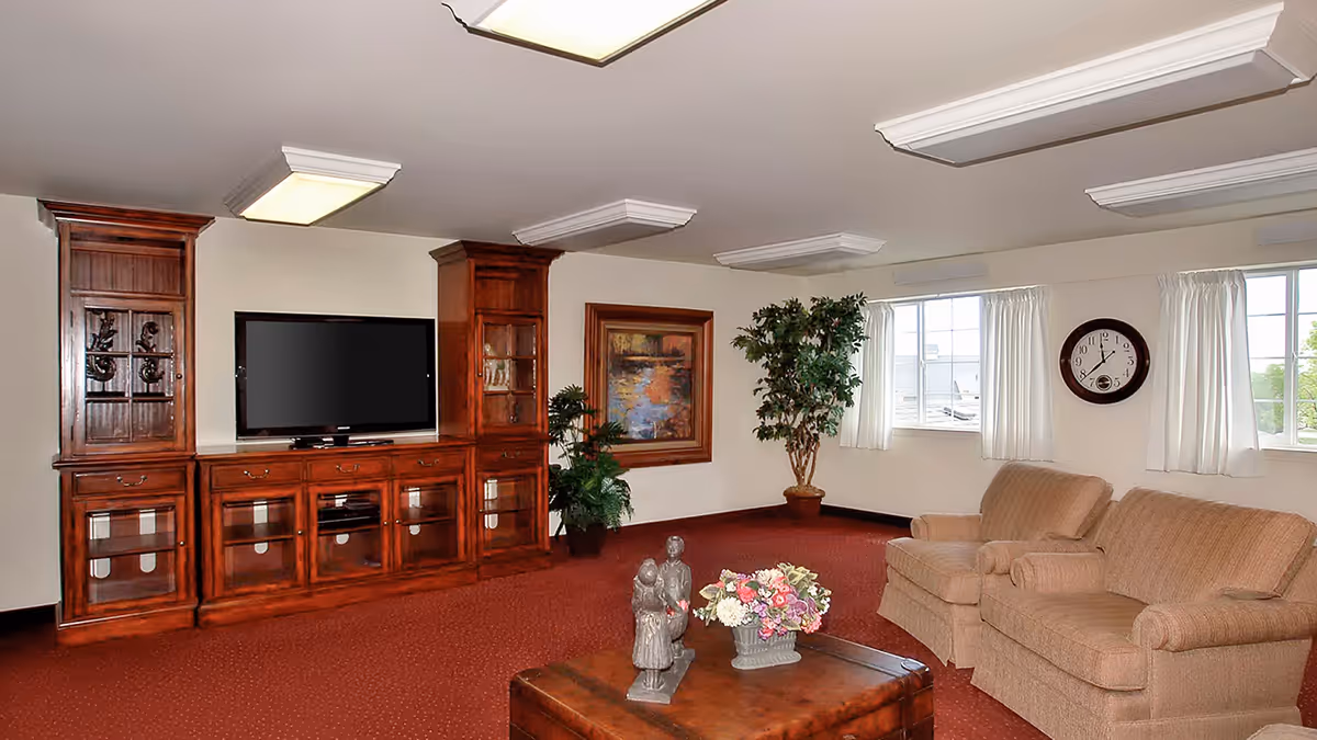 A cozy living room with a large wooden entertainment center holding a flat-screen TV, two beige armchairs, a wooden coffee table with decorative figurines and a flower arrangement, a large framed painting on the wall, two potted plants, a round wall clock, and windows with white curtains letting in natural light.