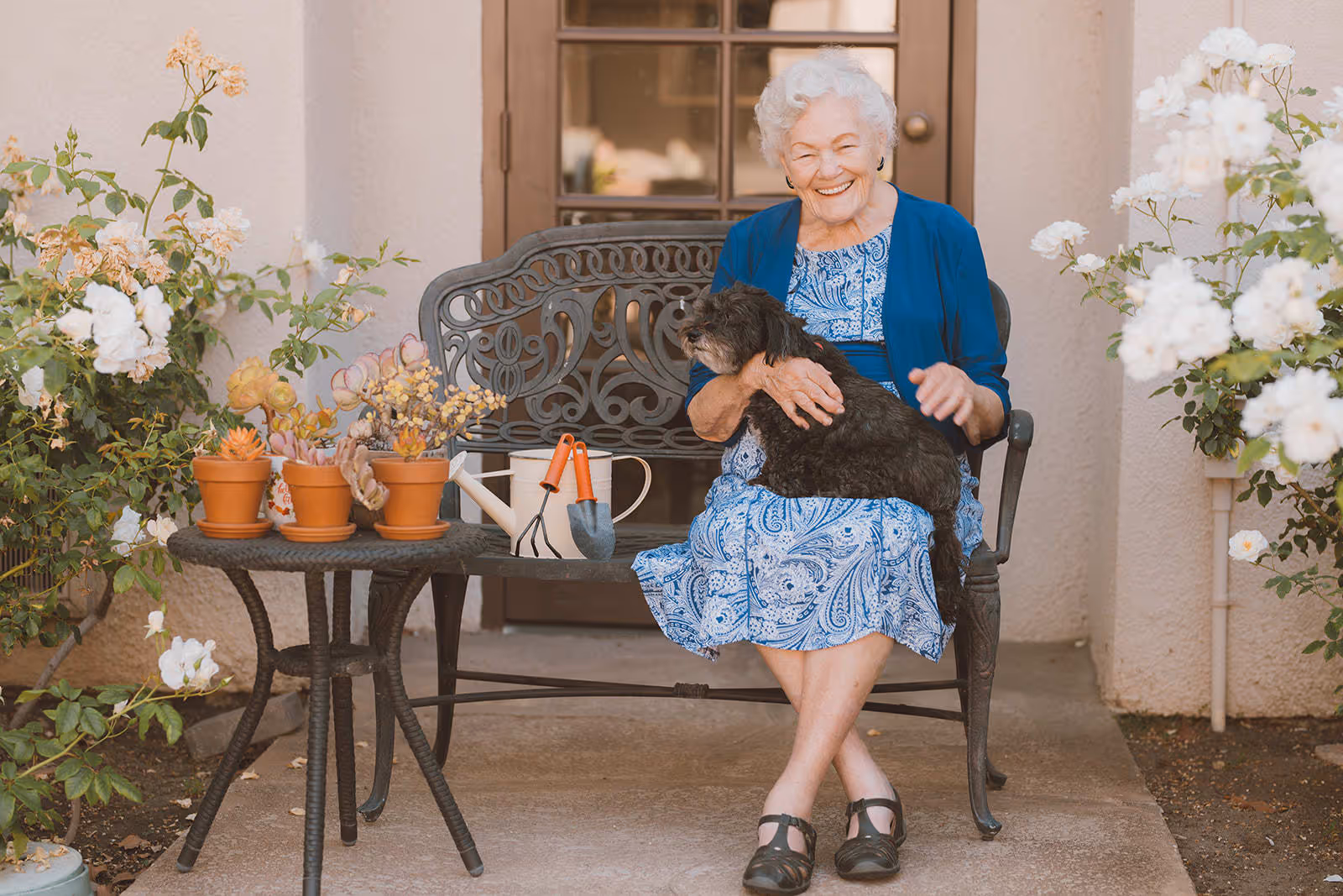 An elderly woman sitting on a decorative metal bench outside, smiling and holding a small black dog on her lap. Next to her is a small table with potted plants, gardening tools, and a watering can. The setting is a garden area with blooming white flowers and a door in the background.