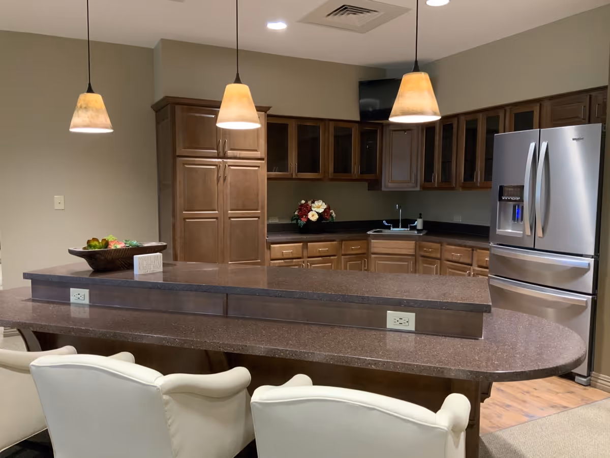 A modern kitchen area with brown wooden cabinets, a stainless steel refrigerator, a double sink, and a dark brown countertop island with three pendant lights hanging above. There are three white cushioned chairs in front of the island and a decorative bowl with fruit on the counter.