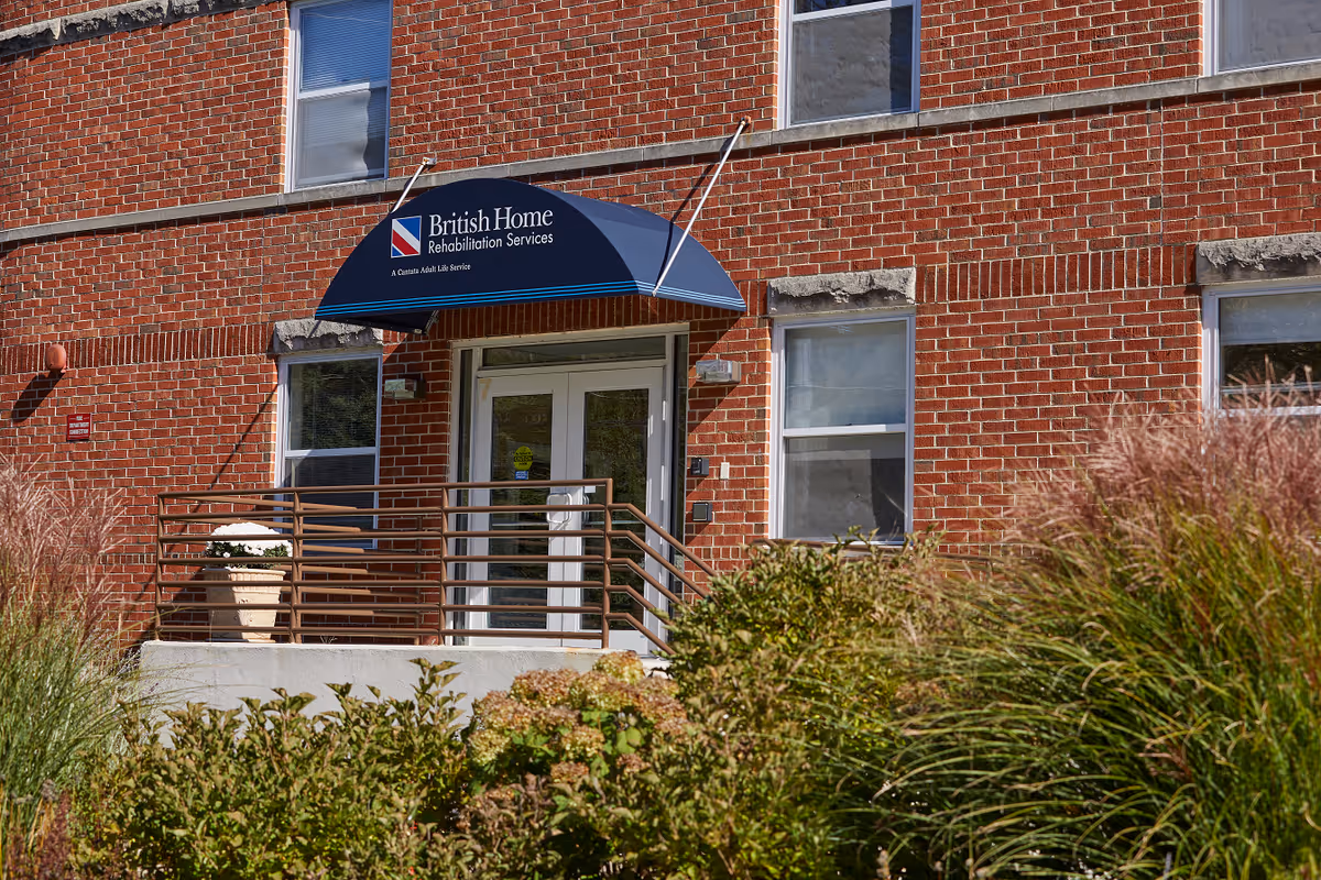 Entrance to a brick building with a blue awning that reads 'British Home Rehabilitation Services, A Cantata Adult Life Service'. There are windows on the building and plants and bushes in the foreground.
