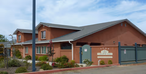 Exterior view of a single-story building with a reddish-brown stucco finish and a gray shingled roof. The building has multiple windows and a gated entrance with a sign that reads 'Lakespur Lodge'. There are small bushes and plants along the front, and a clear blue sky with some clouds above.