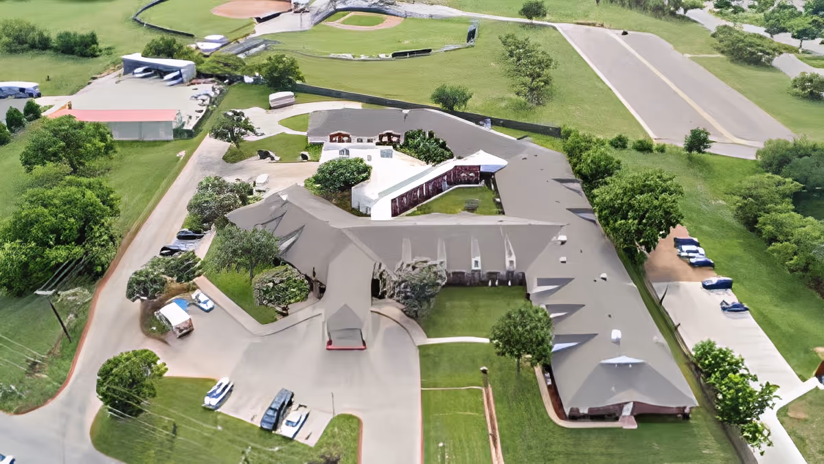 Aerial view of a senior living facility named Morada Abilene, showing a large building with a gray roof surrounded by green lawns, trees, parking areas, and nearby sports fields including a baseball diamond.
