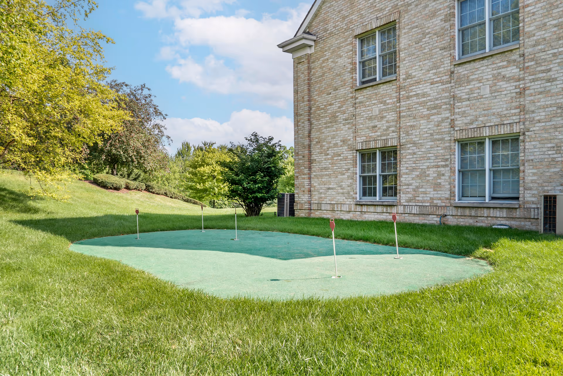 Outdoor putting green with five small flags on a grassy lawn next to a brick building under a partly cloudy sky.