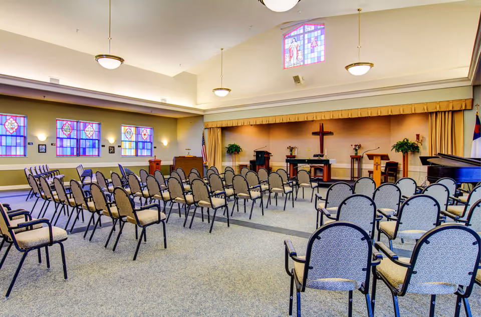 A spacious chapel or meeting room with rows of cushioned chairs arranged facing a stage area. The stage features a wooden cross, podiums, plants, and a piano on the right side. The room has stained glass windows on the left wall and a high ceiling with hanging light fixtures.