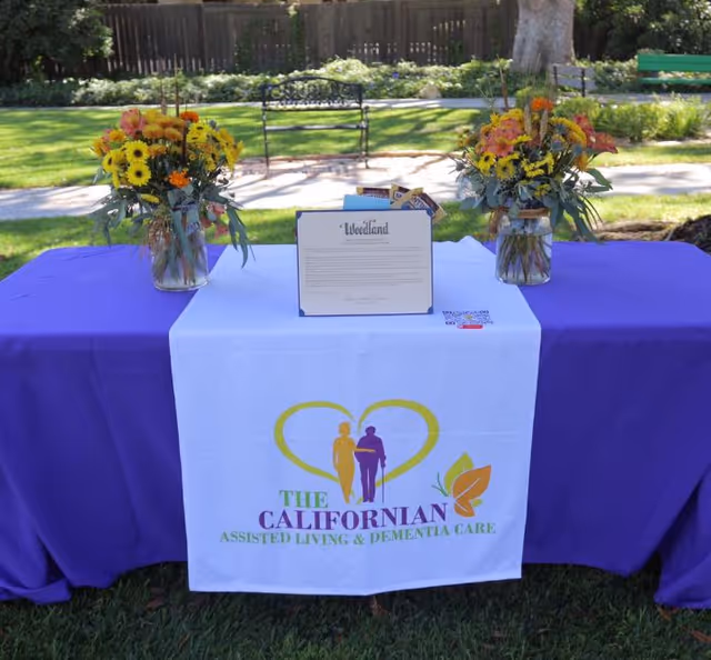 Outdoor table with a purple cloth and a white banner reading 'The Californian Assisted Living & Dementia Care', flanked by two vases of flowers and a framed sign on a grassy park lawn.