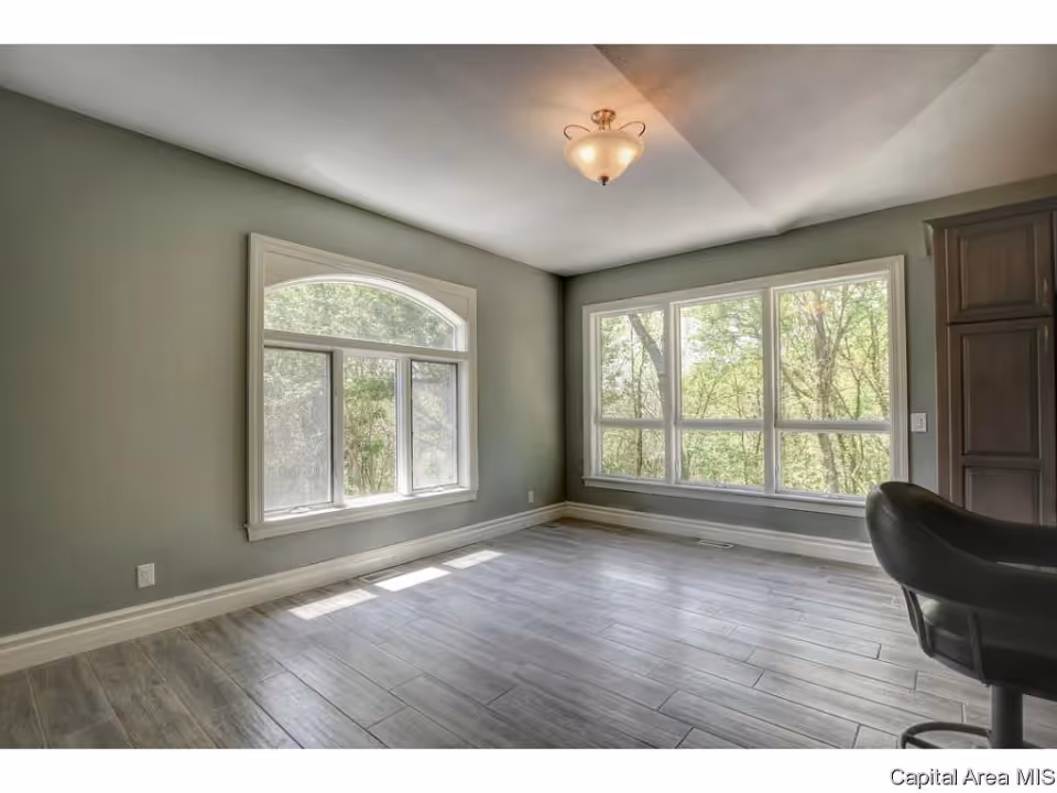 Empty bright room with large windows, gray-green walls, wood-look tile floor, and a ceiling light.