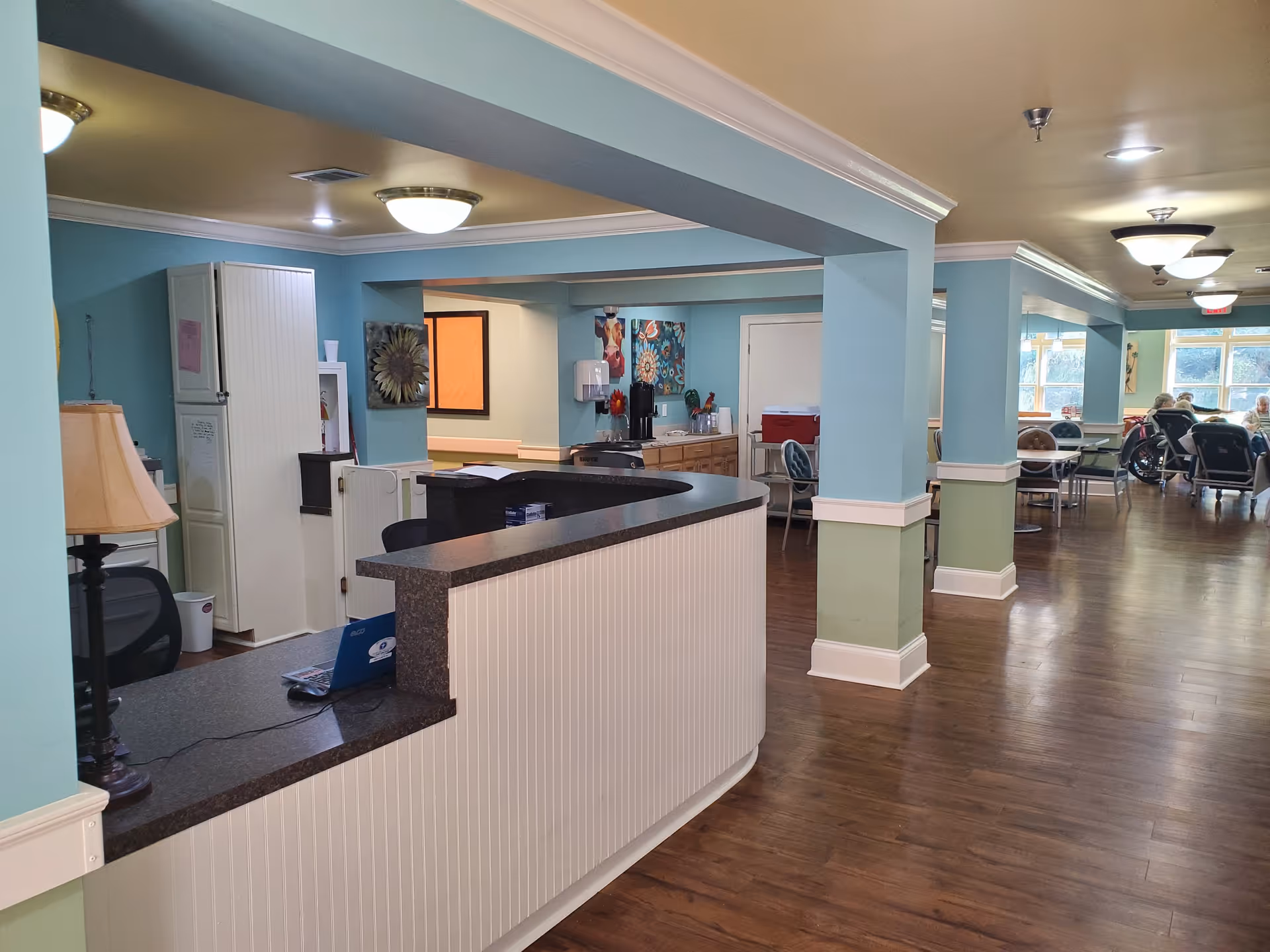 Interior view of a senior living facility reception area with a curved counter and a lamp on it. The walls are painted light blue and green with white trim. In the background, there is a dining area with tables and chairs, some occupied by elderly residents, including one in a wheelchair. The floor is wooden, and ceiling lights illuminate the space.