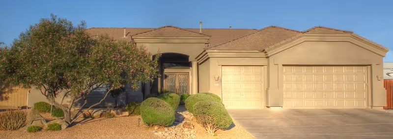 Front exterior of a single-story stucco house with a three-car garage, driveway and desert landscaping.