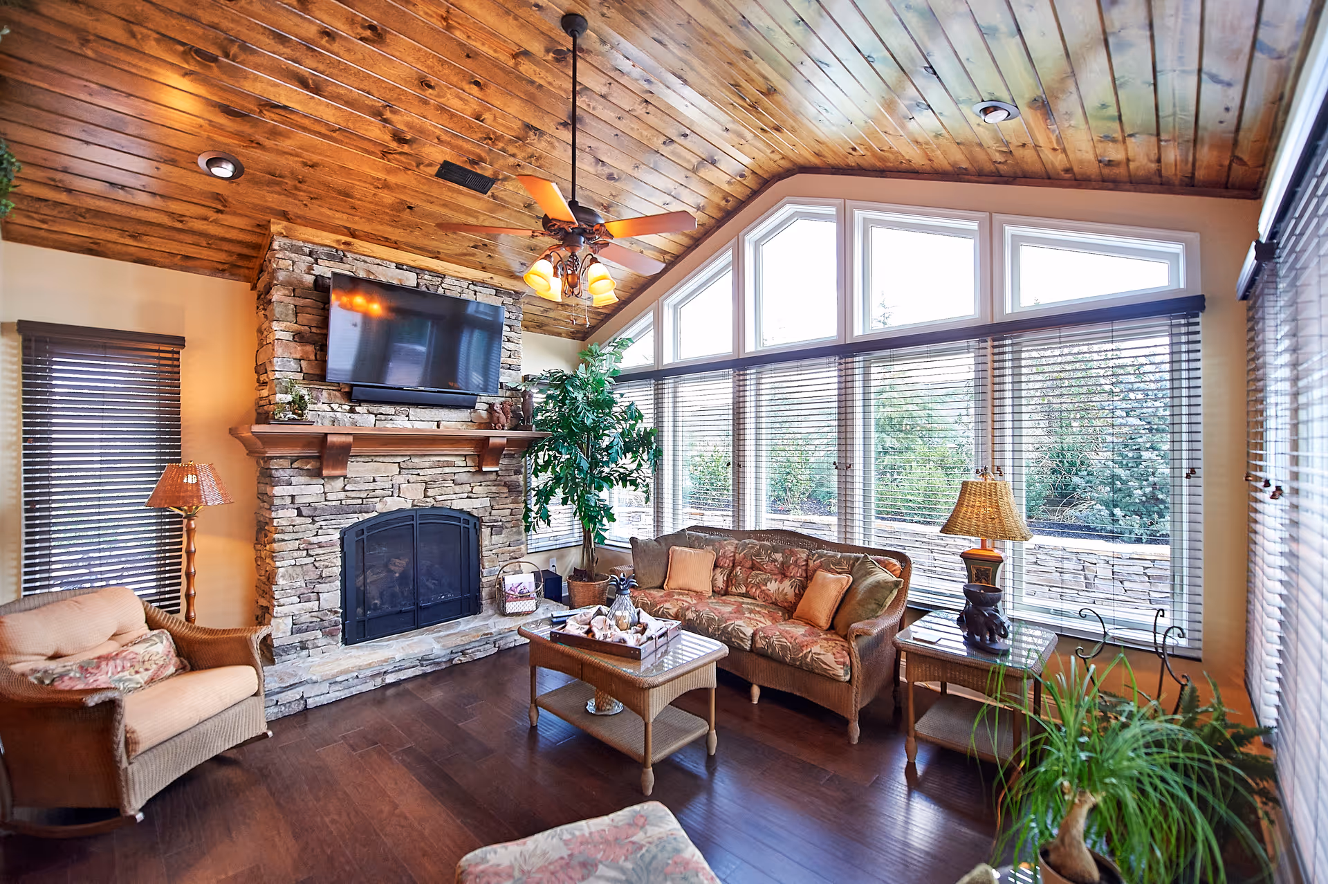 A cozy living room with a wooden ceiling and dark hardwood floor. The room features a stone fireplace with a mounted flat-screen TV above it, a ceiling fan with lights, large windows with blinds letting in natural light, a floral-patterned sofa, a wicker armchair, a coffee table with decorative items, side tables with lamps, and several potted plants.