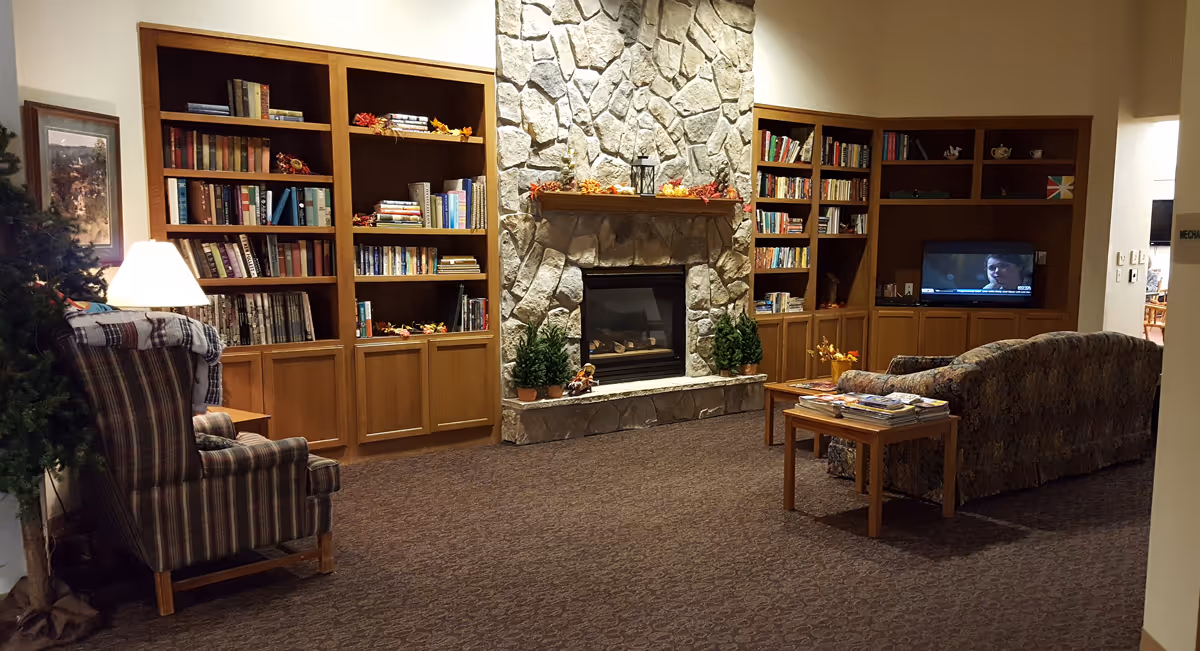 A cozy lobby featuring a stone fireplace, bookshelves filled with books, and comfortable seating. A small table holds magazines, and a television is visible in the background.
