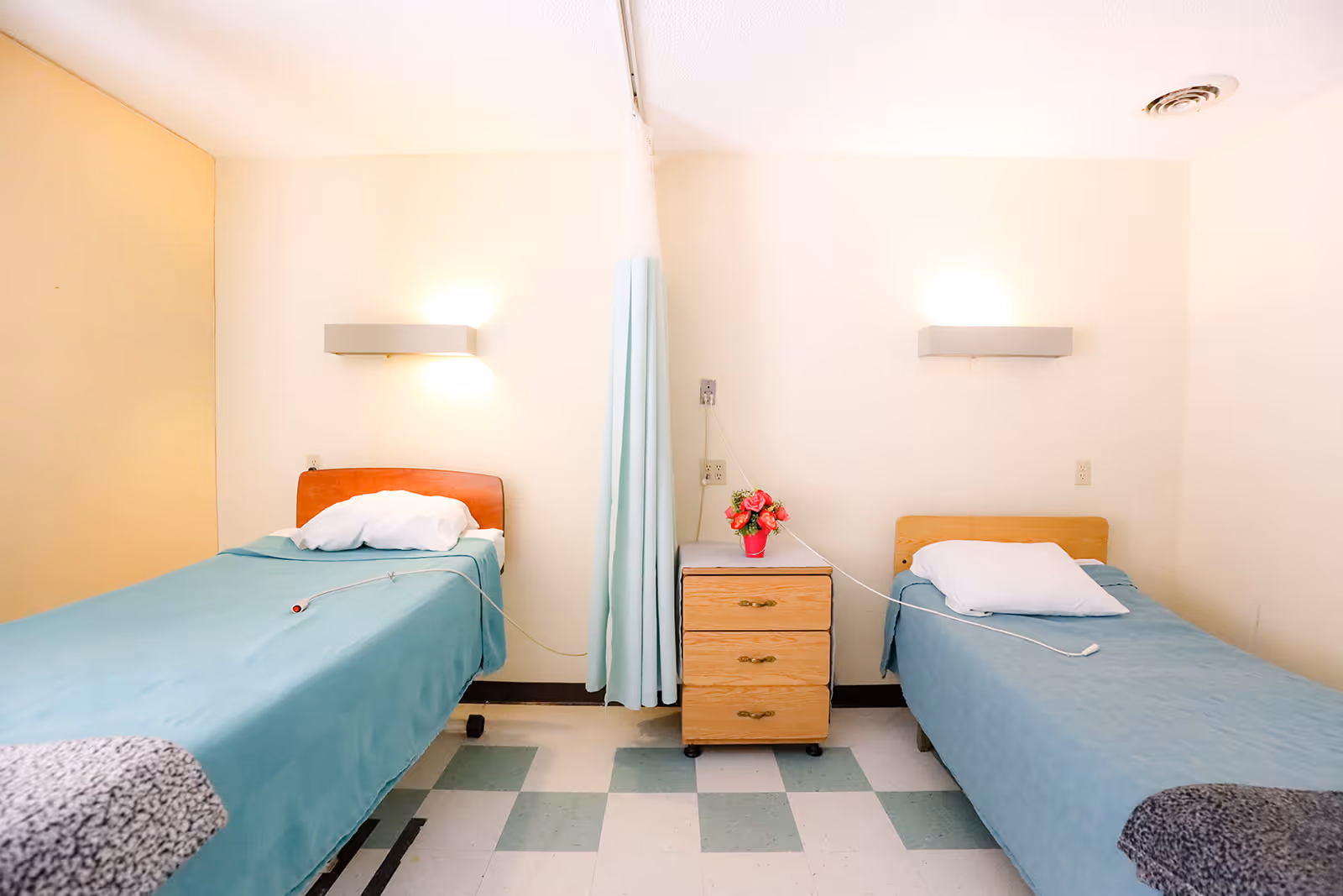 A simple room with two single beds separated by a small wooden nightstand with three drawers and a small vase of pink flowers on top. Each bed has a blue bedspread, a white pillow, and a call button cord. A light fixture is mounted on the wall above each bed, and a light blue privacy curtain hangs between the beds. The floor has a checkered pattern with green and white tiles.