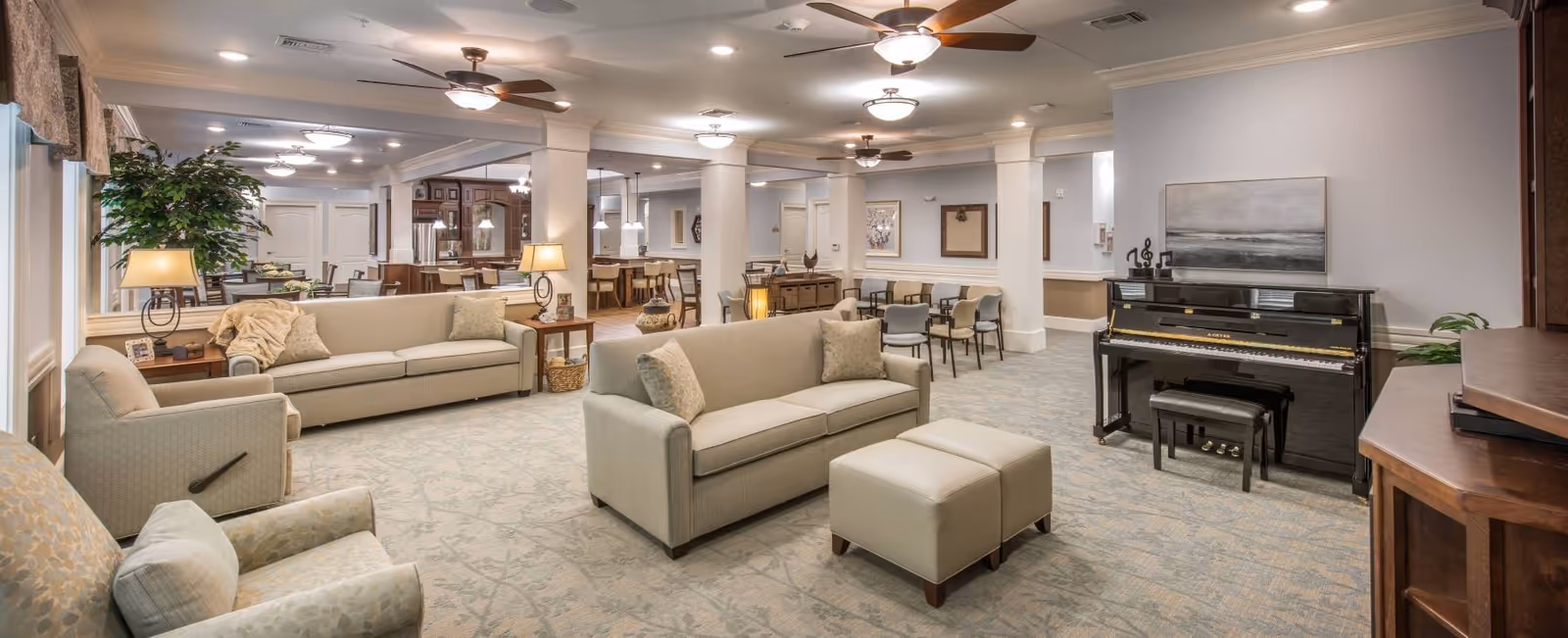 A spacious and well-lit senior living common area featuring beige sofas, armchairs, a piano with a bench, ceiling fans, and soft lighting. The room has a carpeted floor with a subtle pattern, light-colored walls, and several columns. In the background, there is a dining area with tables and chairs, and a kitchen area with wooden cabinetry and pendant lights.