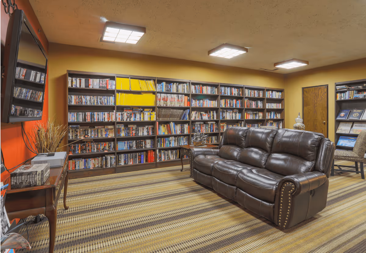 A cozy library or reading room with a large brown leather sofa, multiple bookshelves filled with books and magazines, a flat-screen TV mounted on an orange wall, and a striped carpeted floor. The room has warm yellow walls and ceiling lights.