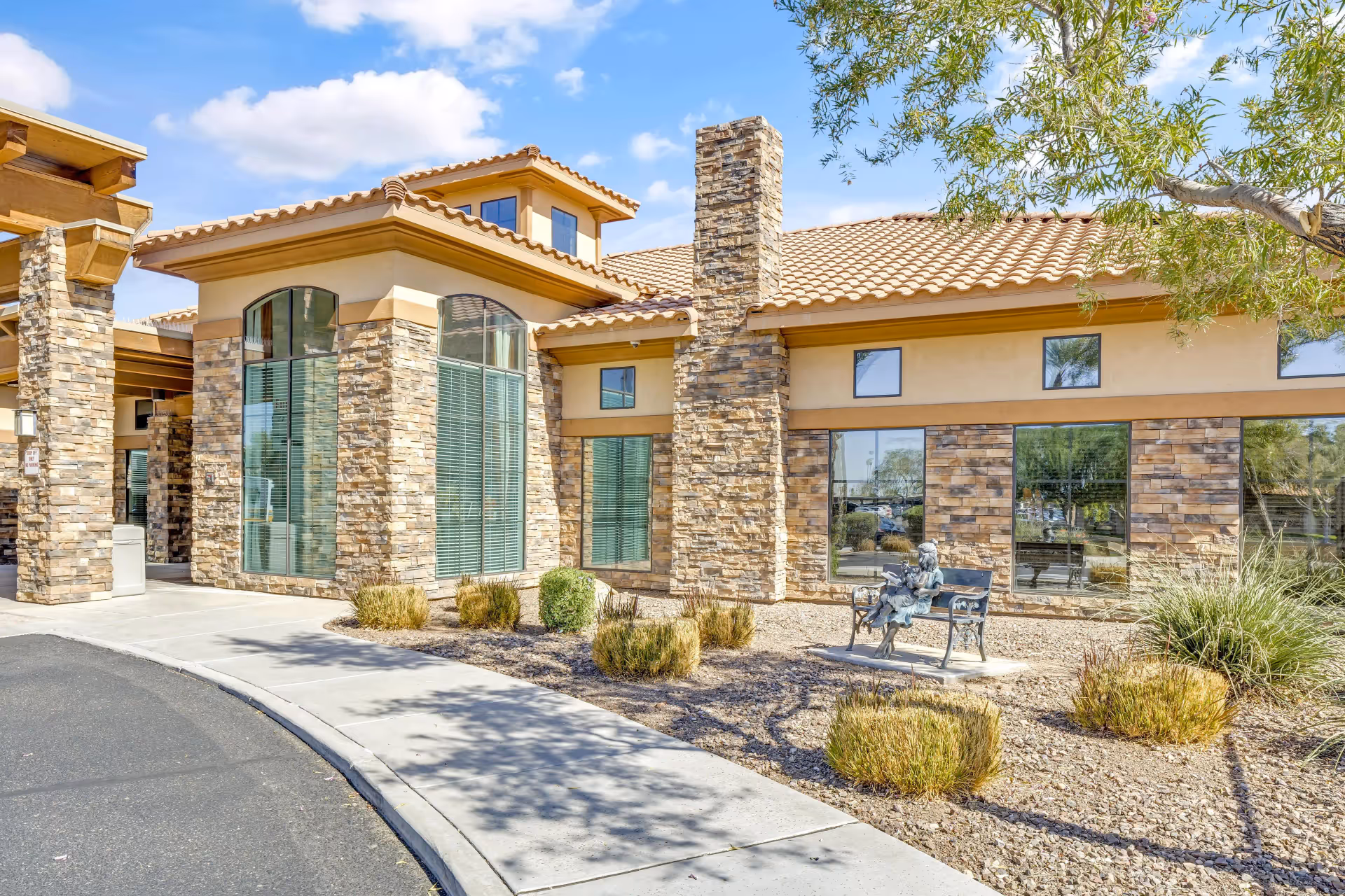 Stone-and-stucco senior living facility front with large windows, landscaped gravel beds, and a bench sculpture under a blue sky.