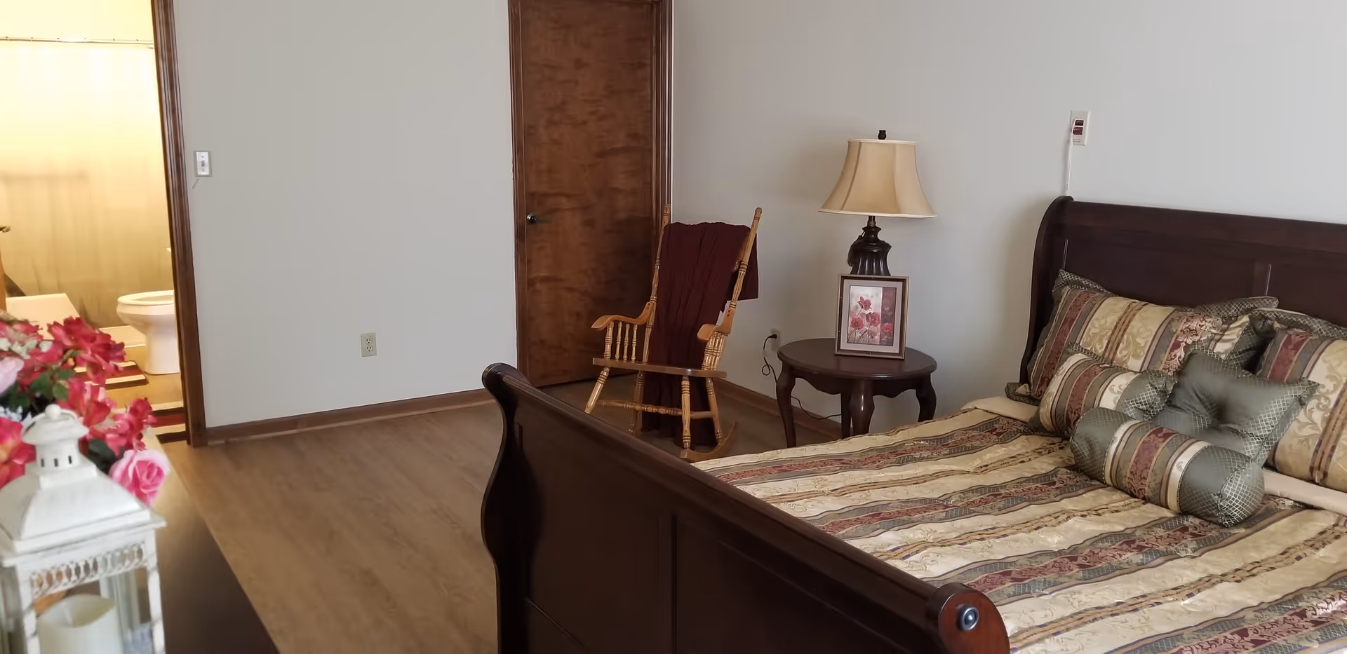 A furnished bedroom with a wooden sleigh bed and patterned bedding, a nightstand with a lamp and framed photo, a rocking chair, and an open doorway revealing a bathroom.