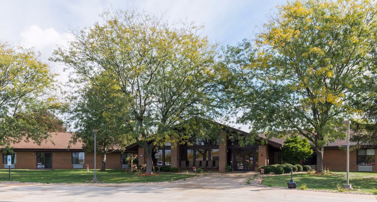 Exterior view of The Loft Rehabilitation and Nursing of Canton building with a brown roof, large windows, and surrounded by green trees and a well-maintained lawn under a partly cloudy sky.