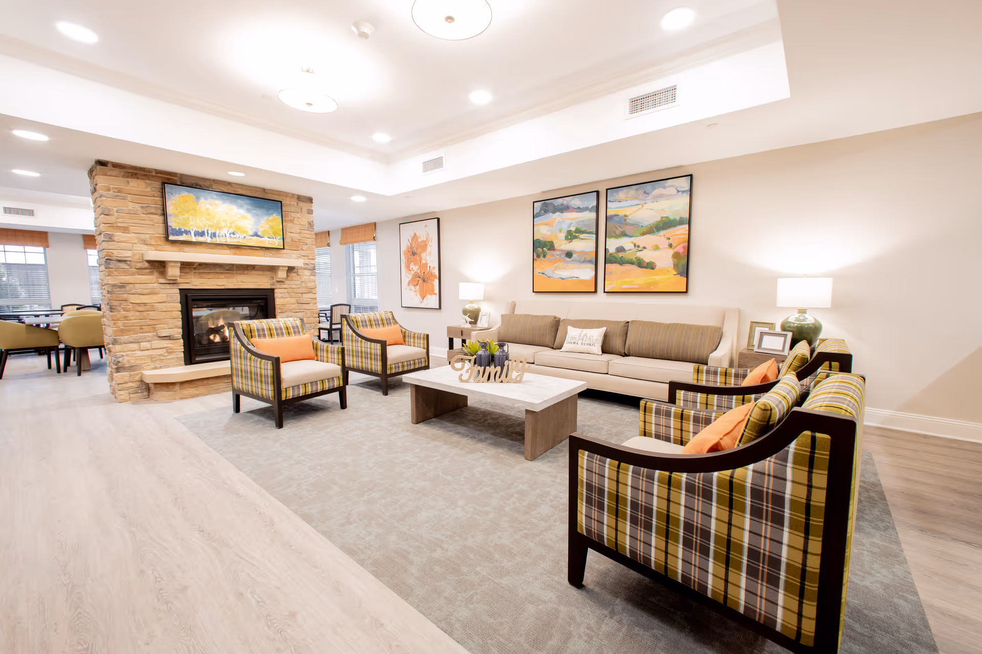 A bright and spacious senior living facility common area featuring a stone fireplace with a painting above it, a beige sofa, four plaid armchairs with orange cushions, a rectangular coffee table with decorative items, two side tables with lamps, and colorful landscape paintings on the wall.