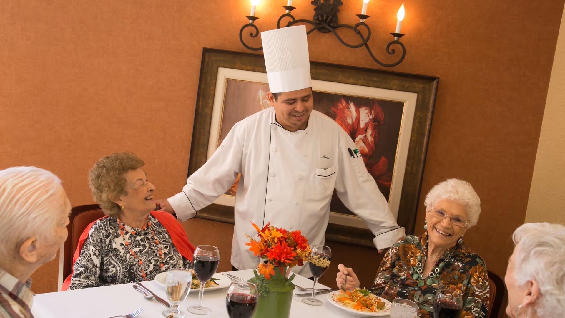 A chef in a white uniform and tall hat stands smiling and interacting with four elderly people seated around a dining table set with plates of food, glasses of wine, and a floral centerpiece in a warmly decorated room.