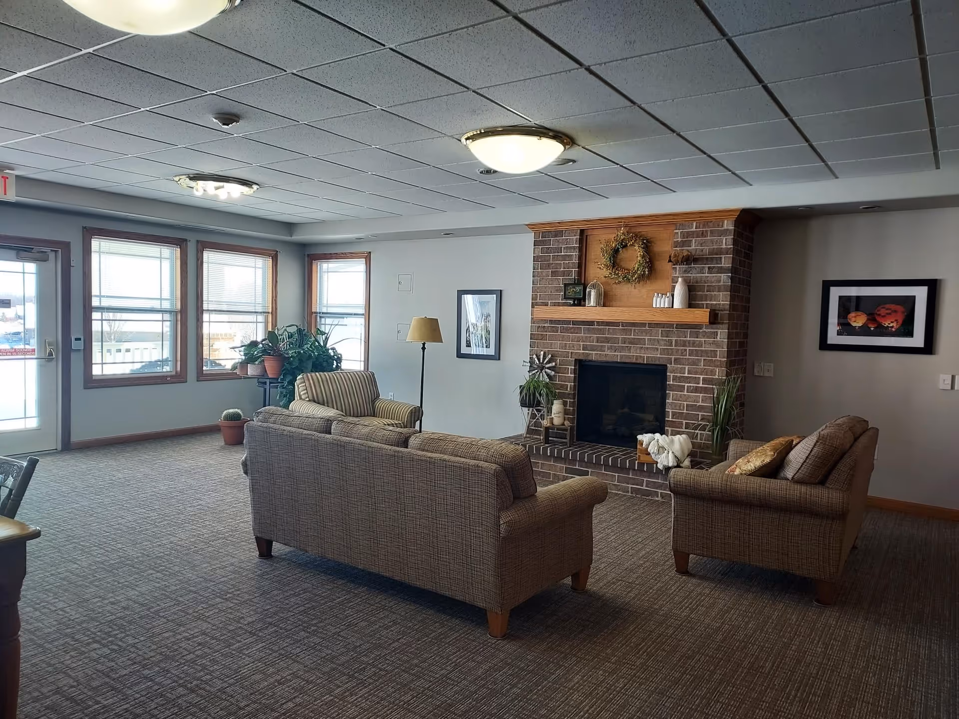 A cozy living room area with two patterned sofas and an armchair arranged around a brick fireplace. The fireplace mantel is decorated with a wreath and various small decorative items. There are framed pictures on the walls, a floor lamp, and several potted plants near windows that let in natural light. The room has a carpeted floor and a drop ceiling with ceiling lights.