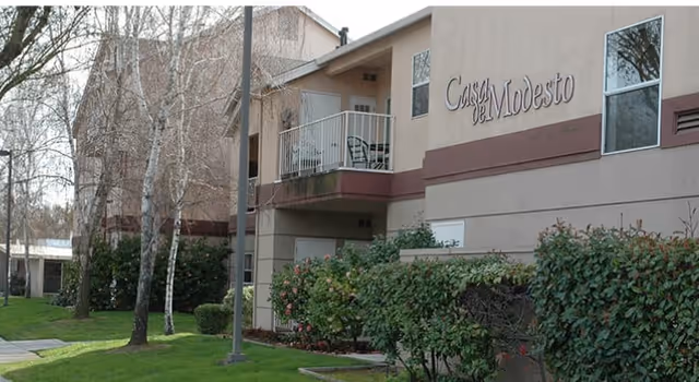 Exterior view of a two-story residential building with beige and brown walls, featuring a balcony with chairs. The building has the name 'Casa de Modesto' displayed on the upper right side. There are trees, bushes, and a well-maintained lawn in the foreground.